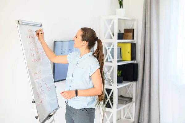 Eine junge Frau im schicken lässigen Hemd schreibt auf Flipchart und lächelt. Seitenansicht — Stockfoto