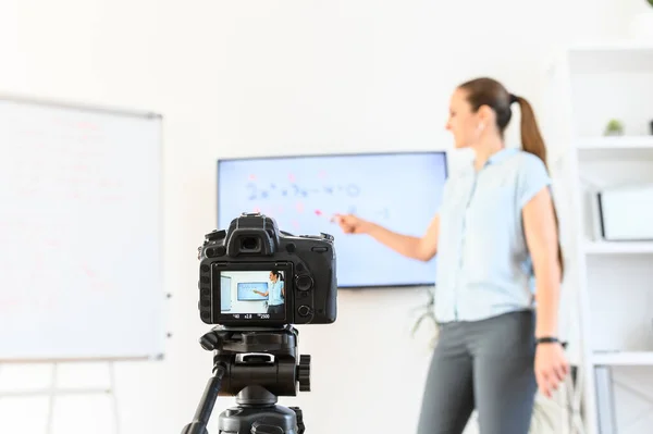 Étude en ligne, apprentissage en ligne. Enseignante enregistre des cours sur une caméra, elle se tient près du tableau à feuilles mobiles et de l'écran avec un marqueur à la main, caméra au premier plan — Photo