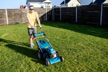 Genç bir adam çim biçme makinesiyle çim biçiyor.