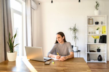 Çevrimiçi öğrenme kavramı, eğitici webinarlar. Genç bir kadın elinde dizüstü bilgisayarla masada oturup internetten video dersleri izliyor. Not defterine bir şey yazmış.