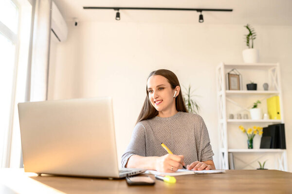 Concept of online learning, educational webinars. A young woman sits at a table with a laptop and watches video lessons online. She notes something in notebook