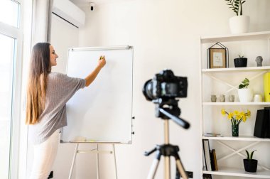 Genç bir kadın internetten yabancı dil öğrenme videosu kaydediyor. Kapalı bir yerde elinde bir çizelgeyle duruyor, üzerine yazıyor ve bir şeyler anlatıyor, kamera üç ayaklı video kaydediyor.