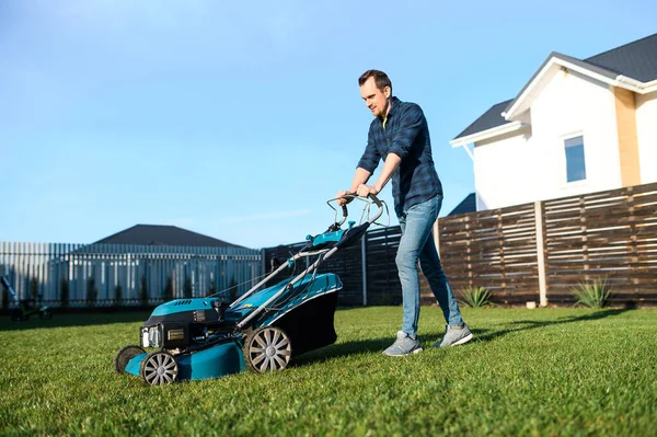 Young man in a plaid shirt and jeans mows lawn — Stock Photo, Image