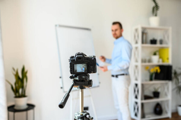 Young male tutor on the camera screen. A guy in smart casual stands near flipcharta in a bright room and records himself on camera. Concept of tutorial videos