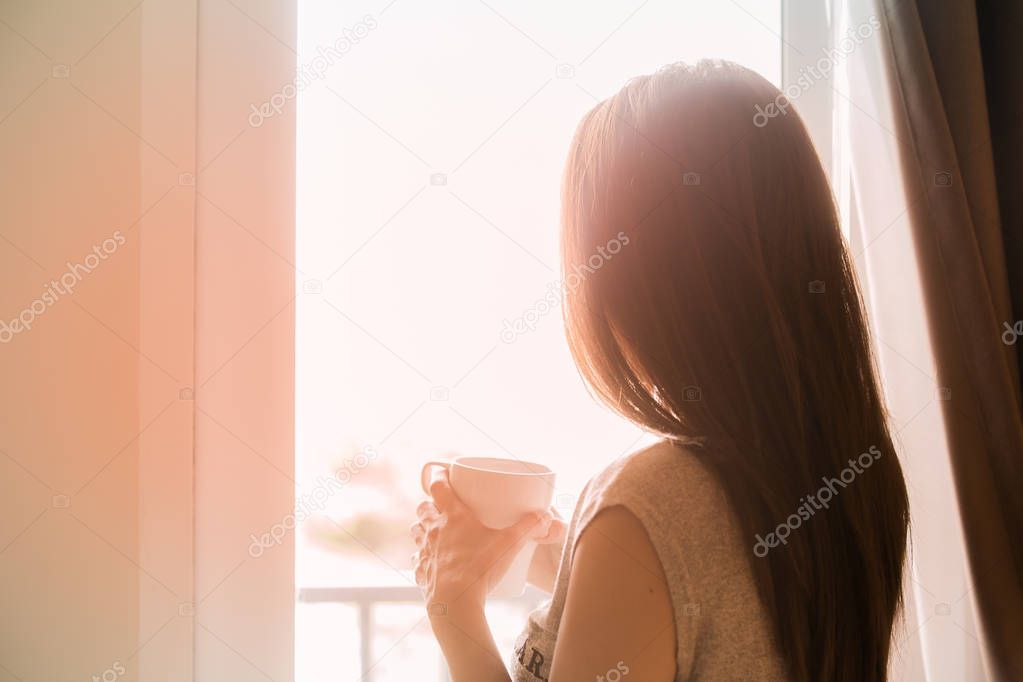 Standing woman holding coffee cup looking out the window.