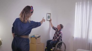 Tekerlekli sandalyedeki engelli koca çerçeveli resim için beyaz bir duvar seçerken, sevimli karısı da çerçeveyi parmaklarından, iç kısımlarından yapıyor.