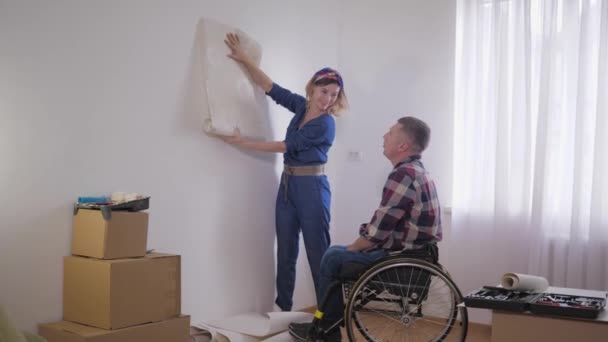 Relaciones familiares, amado hombre discapacitado en silla de ruedas con el cuidado, esposa sonriente elige el papel pintado en el nuevo interior durante la reparación — Vídeo de stock