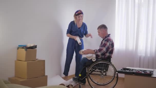 Reparación familiar, amado hombre discapacitado en silla de ruedas con joven hermosa esposa elige el papel pintado en el interior del dormitorio nuevo durante la reparación — Vídeo de stock