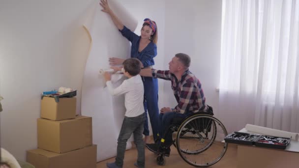 Reparación familiar, padre en silla de ruedas ayuda a la esposa y el niño a elegir nuevo fondo de pantalla para la habitación durante la reparación — Vídeo de stock