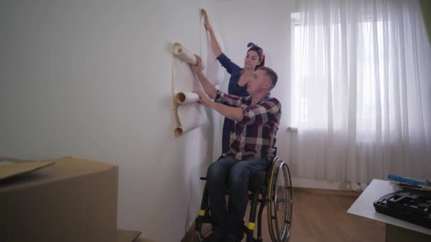 Famille amicale belle jeune femme et heureux mari personne handicapée en fauteuil roulant choisir fond d'écran lors de la réparation dans le nouvel appartement — Video