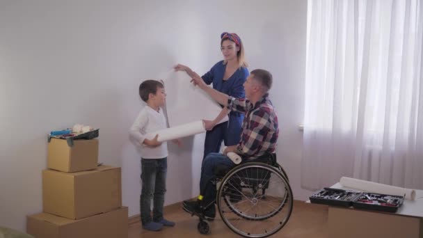 Familia con padre en silla de ruedas elige alegremente nuevo fondo de pantalla en la habitación durante la reparación — Vídeo de stock