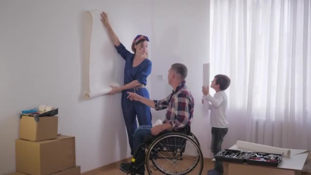 freundliche Familie, geliebter behinderter Vater im Rollstuhl mit fürsorglicher Frau und wunderbarem Kind wählt Tapete in neuem Interieur während der Reparatur