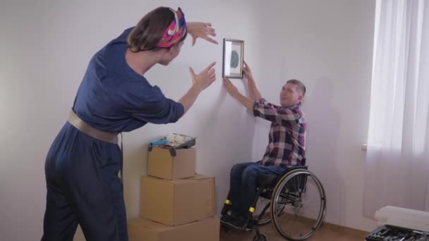 Utile persona disabile in sedia a rotelle appende immagine mentre la moglie fa cornice con le dita e sceglie il posto per l'immagine sulla parete bianca — Video Stock