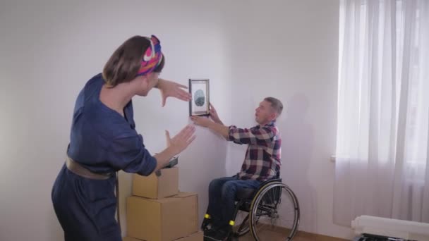 Homme serviable en fauteuil roulant accroche photo tandis que sa femme fait cadre avec ses doigts et choisit un endroit pour une image sur le mur blanc, la décoration d'un appartement — Video