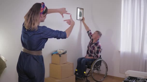 Молодая счастливая пара с мужем в инвалидной коляске переехала в новый дом и повесила декор на белую стену — стоковое видео