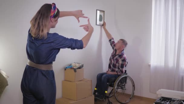 Young happy couple with husband in wheelchair moved to their new home and hung decor on white wall — Stock Footage Young happy couple with husband in wheelchair moved to their new home and hung decor on white wall Stock Video