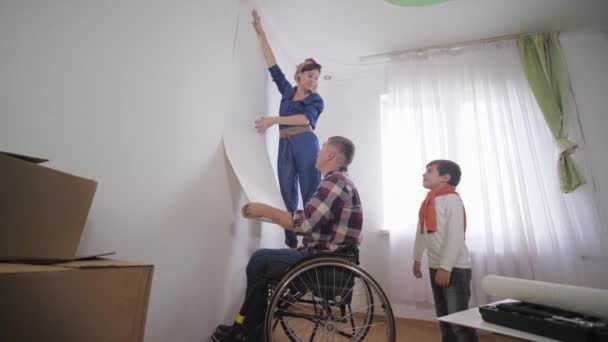 Familia feliz joven padre feliz y persona discapacitada en silla de ruedas con esposa e hijo alegre están eligiendo nuevo fondo de pantalla en la habitación durante la reparación — Vídeos de Stock