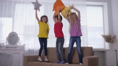 Aktif çocuklar evlerinde tatillerde, ellerinde yastıklarla kanepede, ışık odasının arka planına zıplıyorlar.