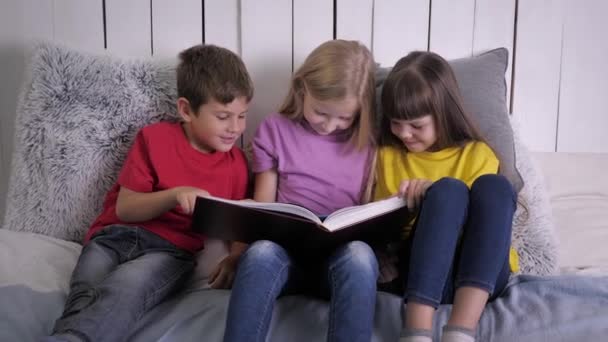 Tres niños inteligentes en ropa multicolor se divierten y leen un libro mientras se sientan en la cama en casa durante las vacaciones — Vídeo de stock