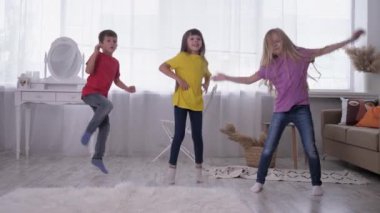 Çok renkli tişörtlerle dans eden çocuklar, beyaz bir odada, beyaz bir pencerenin arkasında, evde tatildeyken eğleniyorlar.