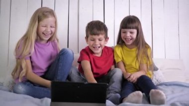 Mutlu çocukluk, neşeli çocuklar dijital tablette komik videolar izliyor ve evde dinlenirken yatakta oturup gülüyorlar.
