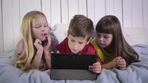 Feliz infancia, los niños amigos en camisetas multicolor juegan en la computadora de la tableta y hablando acostado en una cama en casa — Vídeo de stock
