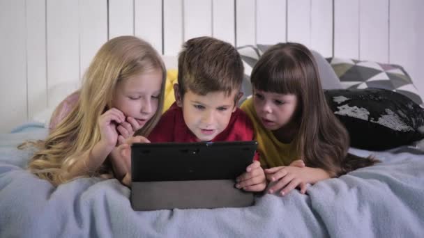 Счастливые маленькие современные друзья в разноцветных футболках используют планшетный компьютер и общаться друг с другом лежа на кровати в квартире — стоковое видео