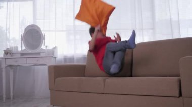 Küçük çocuk kanepede zıplar, çocuk arka planda yastıkla uzanır ve evde ağır çekimde dinlenir.