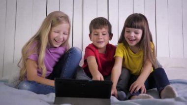 Gülümseyen çocuklar dijital tablette komik videolar izliyorlar evde dinlenirken yatakta oturuyorlar, mutlu bir çocukluk.