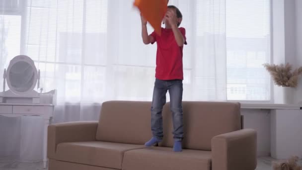 Alegre menino se divertir pulando no sofá e brinca com o travesseiro durante o descanso em casa em câmera lenta — Vídeo de Stock