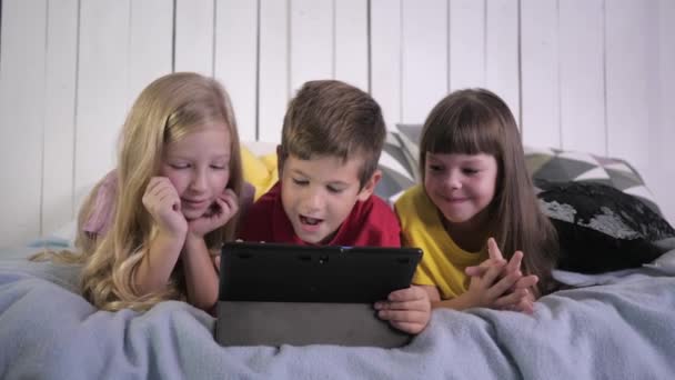 Modern childhood, little friends in multi colored t-shirts use digital tablet and chat while lying on bed at home — Stock Video