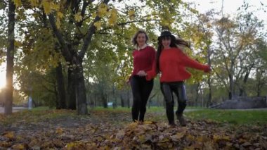 Sonbahar eğlenceleri, gülen genç kadınlar el ele tutuşup sonbahar parkında yürürken sarı yaprakları saçarak eğlenirler.