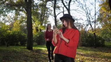 Sonbahar zamanı, çekici, gülümseyen genç kız arkadaşlar akıllı telefondan selfie çeker ve yaprak düşüşü sırasında güneşin altında bir buket sarı yaprak fırlatırlar.