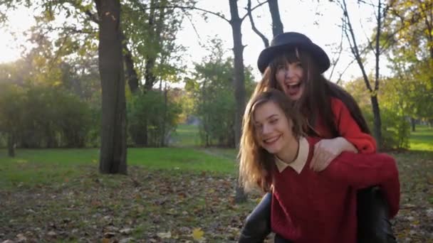 Novias alegres se divierten en el parque de otoño saltando sobre su espalda durante las vacaciones conjuntas en fin de semana soleado — Vídeo de stock