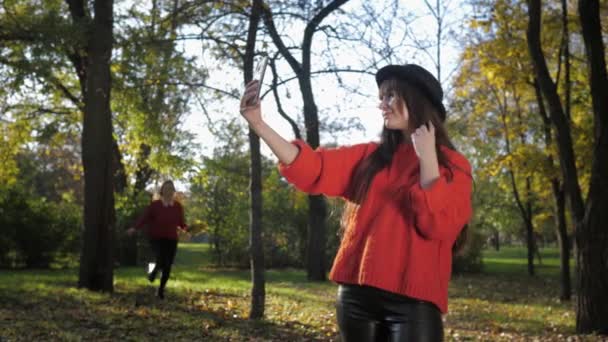Feminidad, chica atractiva joven corre alegremente para conocer a su novia bonita en un sombrero y tomar una selfie en el teléfono con hojas caídas arrojarlo en el hermoso parque — Vídeo de stock