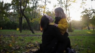 Sonbahar sezonu, yakışıklı kız, bir sonbahar parkında dökülen yaprakları fırlatırken sevgi dolu annenin kollarında oturuyor.