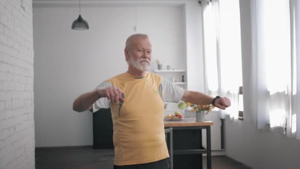 Cuidados de saúde, um homem atraente idoso vai para esportes e executa exercícios para melhorar o corpo, faz gesto impotente e vira de um lado para o outro, enquanto dentro de casa — Vídeo de Stock