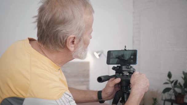 Oudere man en technologie, moderne grootvader is sociaal actief en heeft zijn eigen blog zet camera op smartphone in zijn kamer om nieuwe inhoud op te nemen — Stockvideo