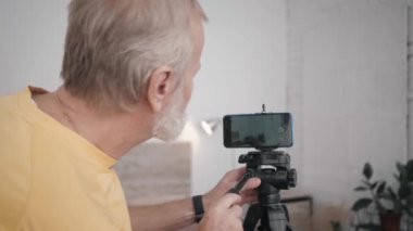 Büyükbaba ve teknoloji, yaşlı erkek blogcu vlog kaydetmek için akıllı telefonunu kullanmaya hazır. Kamerayı kuruyor ve içeriği çekmek için odasına giriyor.