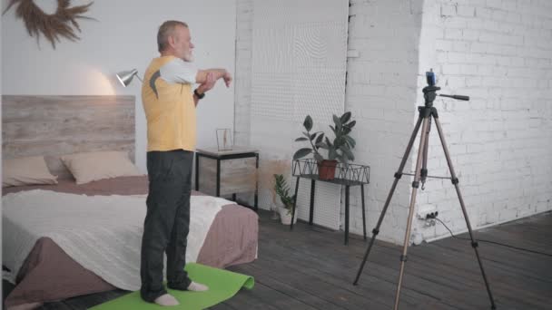 Attrayant vieil homme montre des exercices de bien-être enregistrement smartphone videon pour les réseaux sociaux et les abonnés dans une chambre confortable avec un intérieur design — Video