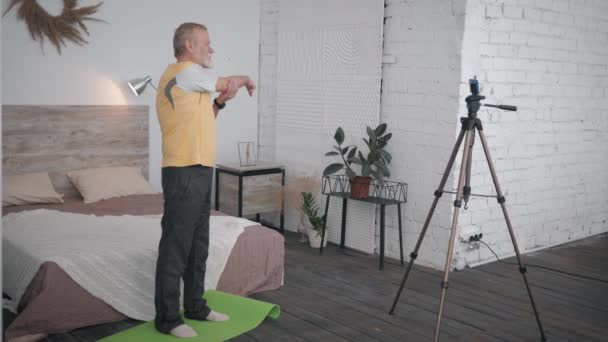 Attraktiv gammal man visar wellness övningar inspelning video smartphone för sociala nätverk och anhängare i mysiga rum med designer interiör — Stockvideo