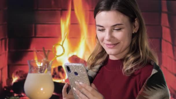 素敵な笑顔の女の子は暖炉の背景にあるエグノグクリスマスカクテルの近くに彼女の手の中に電話で座って、ホームレス — ストック動画