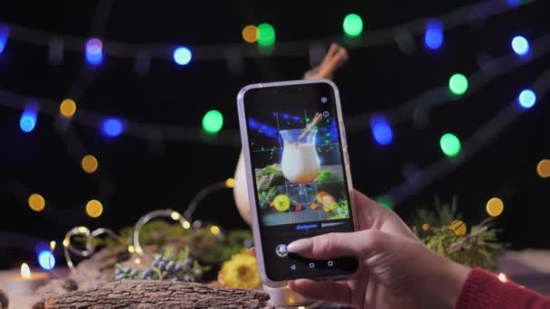 Фотографія дівчини на смартфоні традиційний яєчний коктейль, прикрашений ялинкою та корицею, приготованою вдома для вечірки з друзями на тлі кольорових вогнів — стокове відео