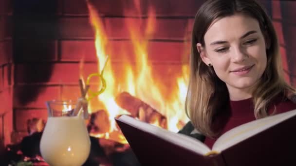 Sıcak, gülümseyen bir kız şöminenin başında oturuyor, bir kitapta ilginç hikayeler okuyor ve arkadaşlarıyla bir parti sırasında evde yapılan geleneksel eggnog kokteylini içiyor. — Stok video