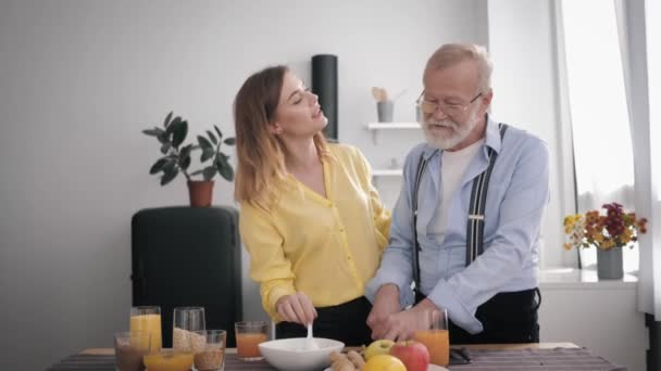 Familj relation, glad pepprig gammal man förbereder en läcker frukost i köket med hälsosamma produkter, blandar med glada vackra vuxna barnbarn vid bordet bakgrund av kitet — Stockvideo