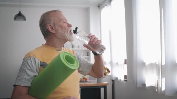 Abuelo deportivo bebe agua limpia para mantener la salud sostiene una estera de yoga en las manos mientras está de pie en el interior con un hermoso interior — Vídeos de Stock