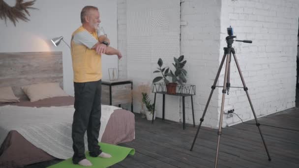 Спортивный блогер, мужчина в возрасте показывает упражнения для суставов на телефоне камера записи видеоблог для последователей в комнате с современным интерьером — стоковое видео