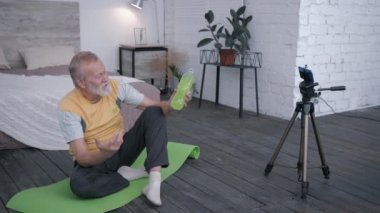 vlogger, sporcu yaşlı blogger video kaydı akıllı telefon blogunda spor içeceğinin video incelemesi abonelerinin yoga minderinin üzerinde oturmasını öneriyor.