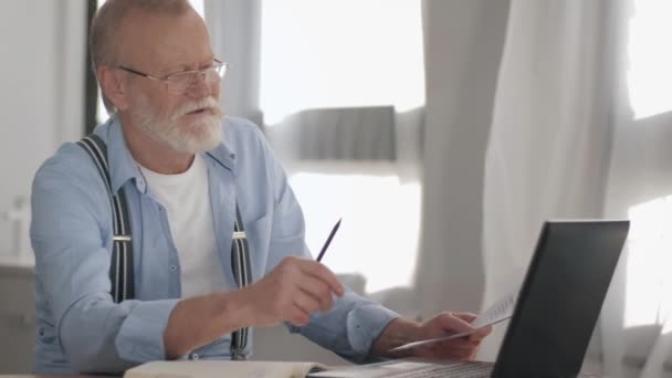 Elderly grandfather with gray beard in glasses for vision works at laptop computer and counts home payments to pay online on computer, portrait of handsome old man — Stockvideo
