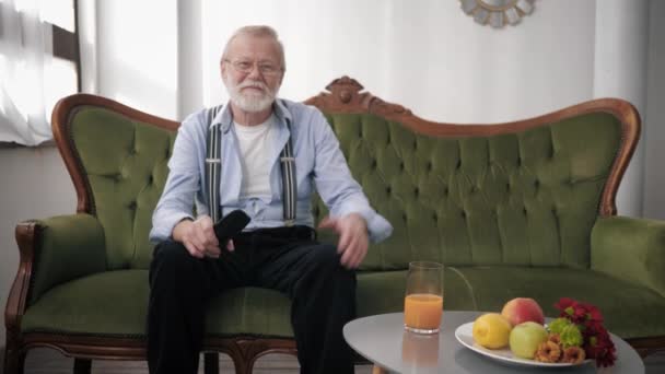 Attraktiver älterer Mann mit Bart und Sehbrille sitzt auf Sofa mit Fernbedienung und verfolgt emotional Fußballspiel — Stockvideo